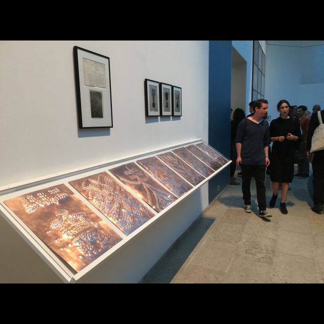 Installation of <i>Sociétés secréts</i>, 2015, at Axis Gallery.<br>
Scarification on 8 bas-relief copper plate, each 29.7 x 42 cm<br>
Fabrication: Dinanderie/Zouak, Morocco<br>
Médailles: Travail et progrès / Médailles: l’union fait la force / Baudouin, Roi des belges. Three black & white photographs.<br>
Secte Punga: Report of a Congolese Detective to the Assistant Director of Kivu (Congo). One hand written letter, 16 x 20 cm, one black-and-white photo 12 x 16 cm. Collection Jean-Pierre Sonck, Brussels