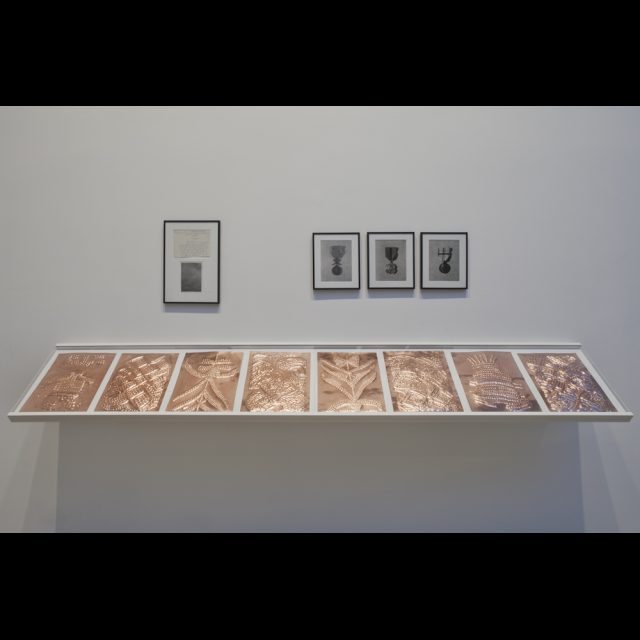 Installation of <i>Sociétés secréts</i>, 2015, at Axis Gallery.<br>
Scarification on 8 bas-relief copper plate, each 29.7 x 42 cm<br>
Fabrication: Dinanderie/Zouak, Morocco<br>
Médailles: Travail et progrès / Médailles: l’union fait la force / Baudouin, Roi des belges. Three black & white photographs.<br>
Secte Punga: Report of a Congolese Detective to the Assistant Director of Kivu (Congo). One hand written letter, 16 x 20 cm, one black-and-white photo 12 x 16 cm. Collection Jean-Pierre Sonck, Brussels