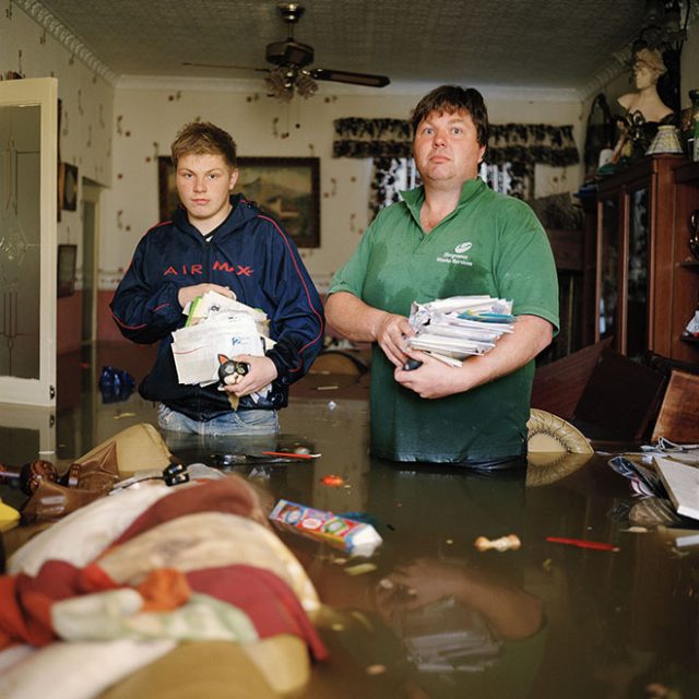 Graham Leith (in green shirt) and his son Kieran in the flooded house of his mother Doreen Leith in Toll Bar village outide Doncaster. Doreen had been too shocked to come back to the house so her son and grandchild had come back to search for her insurance papers. This was one of the communities flooded when a freak storm unleashed a deluge of rain on parts of northern England in which more than four inches of rain fell in 24 hours. The devastation was caused by extreme and unpredictable weather and it is this kind of freak event, which climate scientists have been predicting, will become more frequent as a consequence of global warming.<br>
Chromogenic print on Fuji Crystal Archive<br>
27 1/2” x 27 1/2 / 48” x 48”; Edition: 5 + 2 AP
<br>
70 x 70 cm / 122 x 122 cm; Edition: 3 + 2 AP