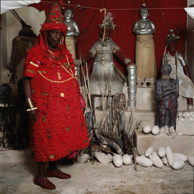 Standing in front of a private shrine whose name means “witches do not speak for God,” this chief wears the hat of his office and the red scalelike tunic representing the pangolin, symbol of chiefly importance. He serves as a symbolic father to the king and as caretaker of the harem, settling disputes between the king’s wives and reporting on their behavior.<br>
Series: <i>Kings, Chiefs, and Women of Power</i><br>
Chromogenic print on Fujiflex Crystal Archive paper<br>
30” x 30”; Edition: 5 + 2 AP<br>
50” x 50”; Edition: 3 + 1 AP