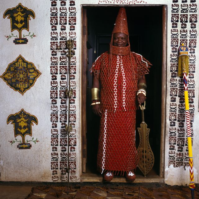 This high-ranking chief wears red-beaded vest, skirt, collar and conical headdress. The ivory bracelets and fish-slice ceremonial sword he holds confirm his political and military status, as does the beaded staff of office resting against the wall.<br>
Series: <i>Kings, Chiefs, and Women of Power</i><br>
Chromogenic print on Fujiflex Crystal Archive paper<br>
30” x 30”; Edition: 5 + 2 AP<br>
50” x 50”; Edition: 3 + 1 AP